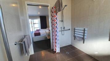 Un baño moderno con ducha, toallero y una cama visible al fondo.