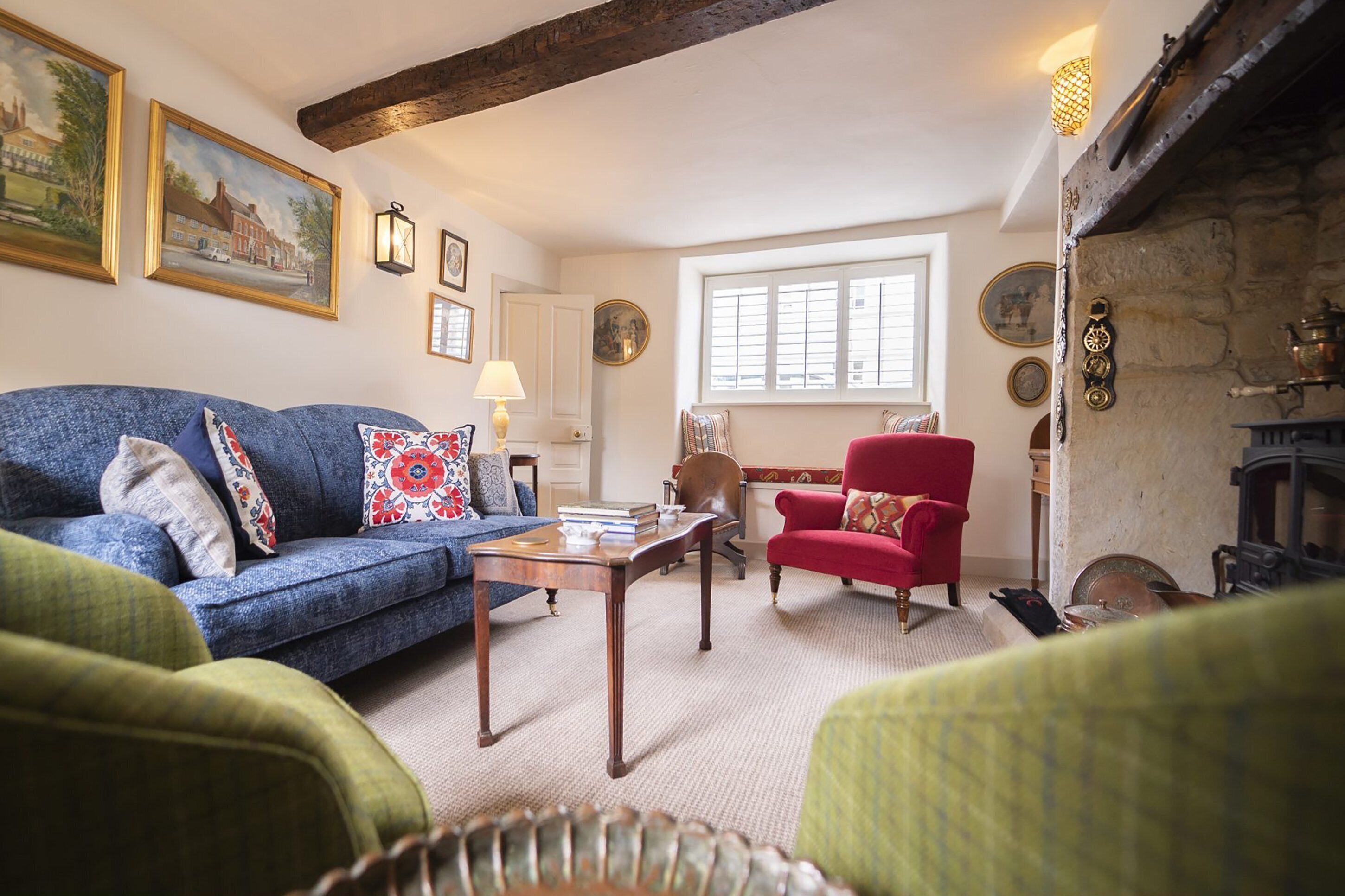 A cozy living room with a blue sofa, a red armchair, and a green armchair. There is a fireplace with a stone mantle and a window with shutters.