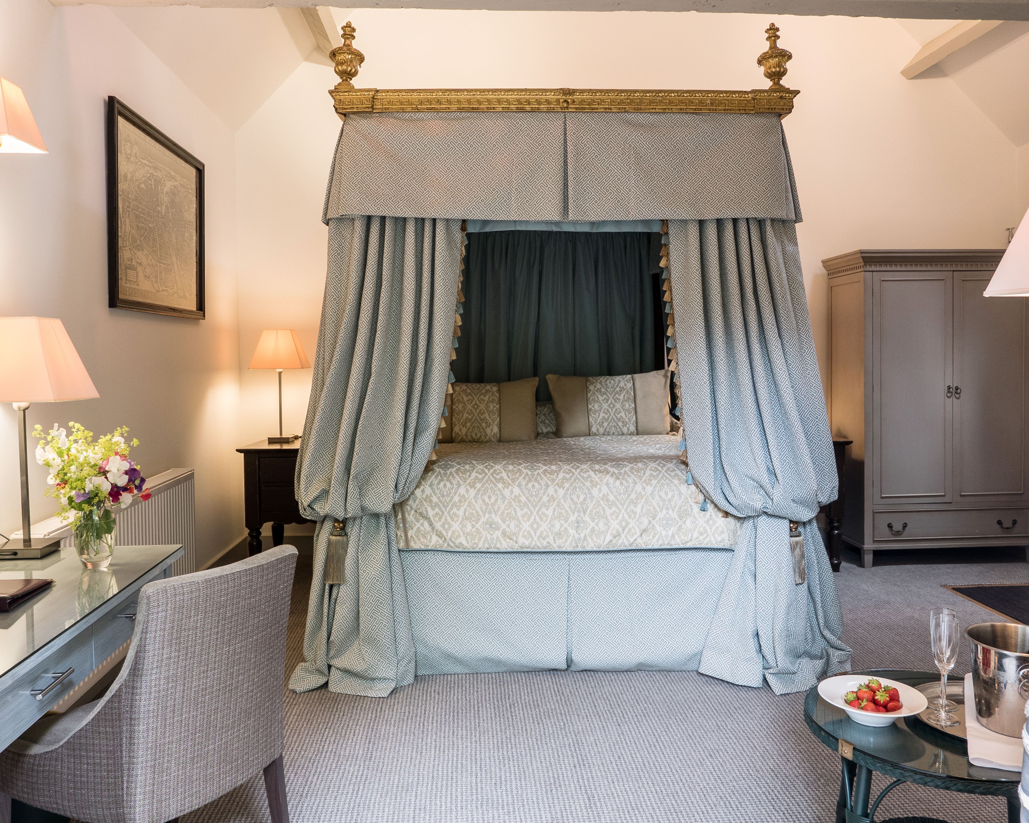 A four-poster canopy bed with a patterned bedspread, a bedside table with a lamp, a chair, and a small table with a bowl of strawberries and a glass.
