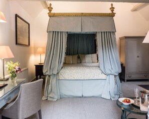 A four-poster canopy bed with a patterned bedspread, a bedside table with a lamp, a chair, and a small table with a bowl of strawberries and a glass.