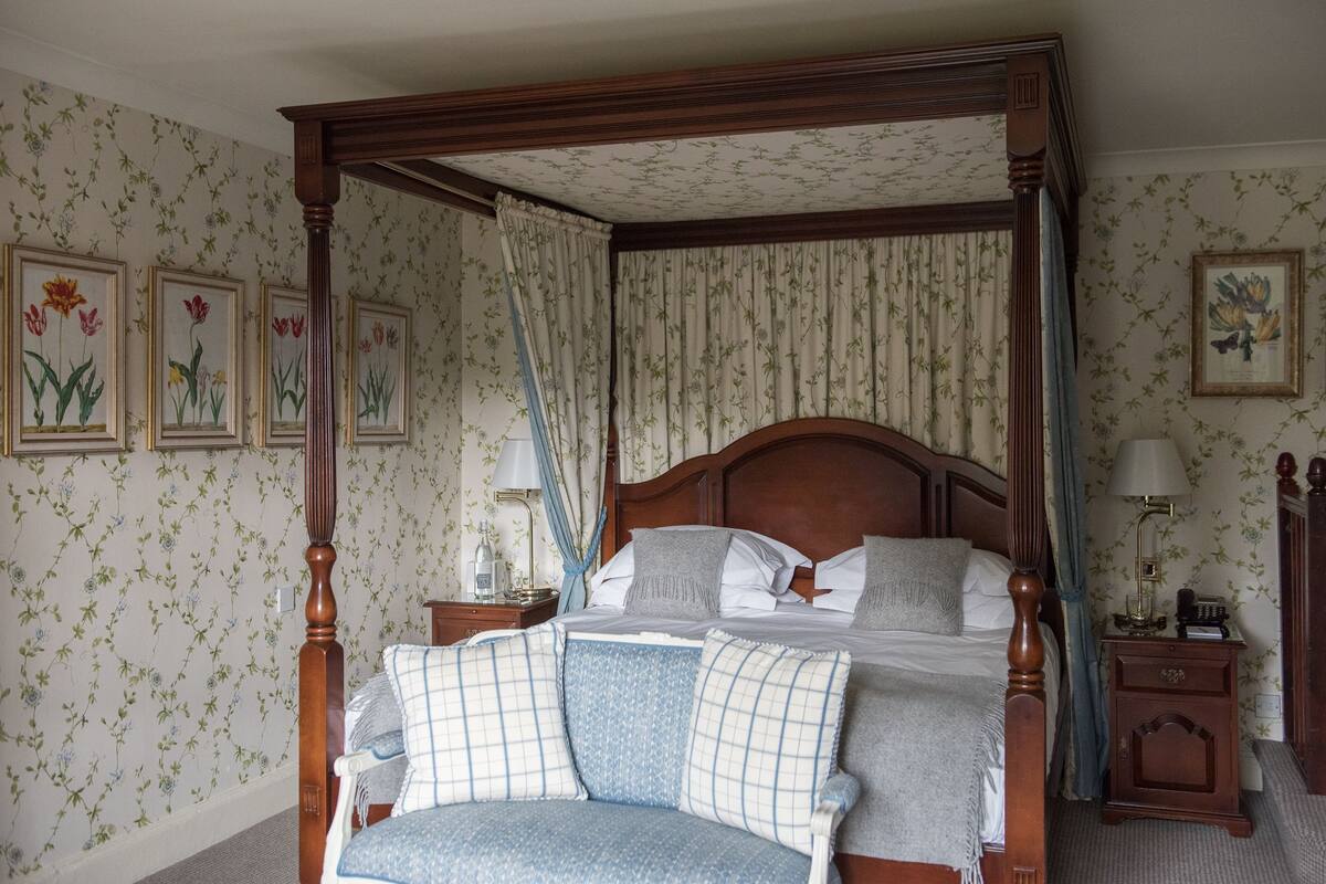 A four-poster bed with a wooden headboard, a nightstand with a lamp, and floral wallpaper.