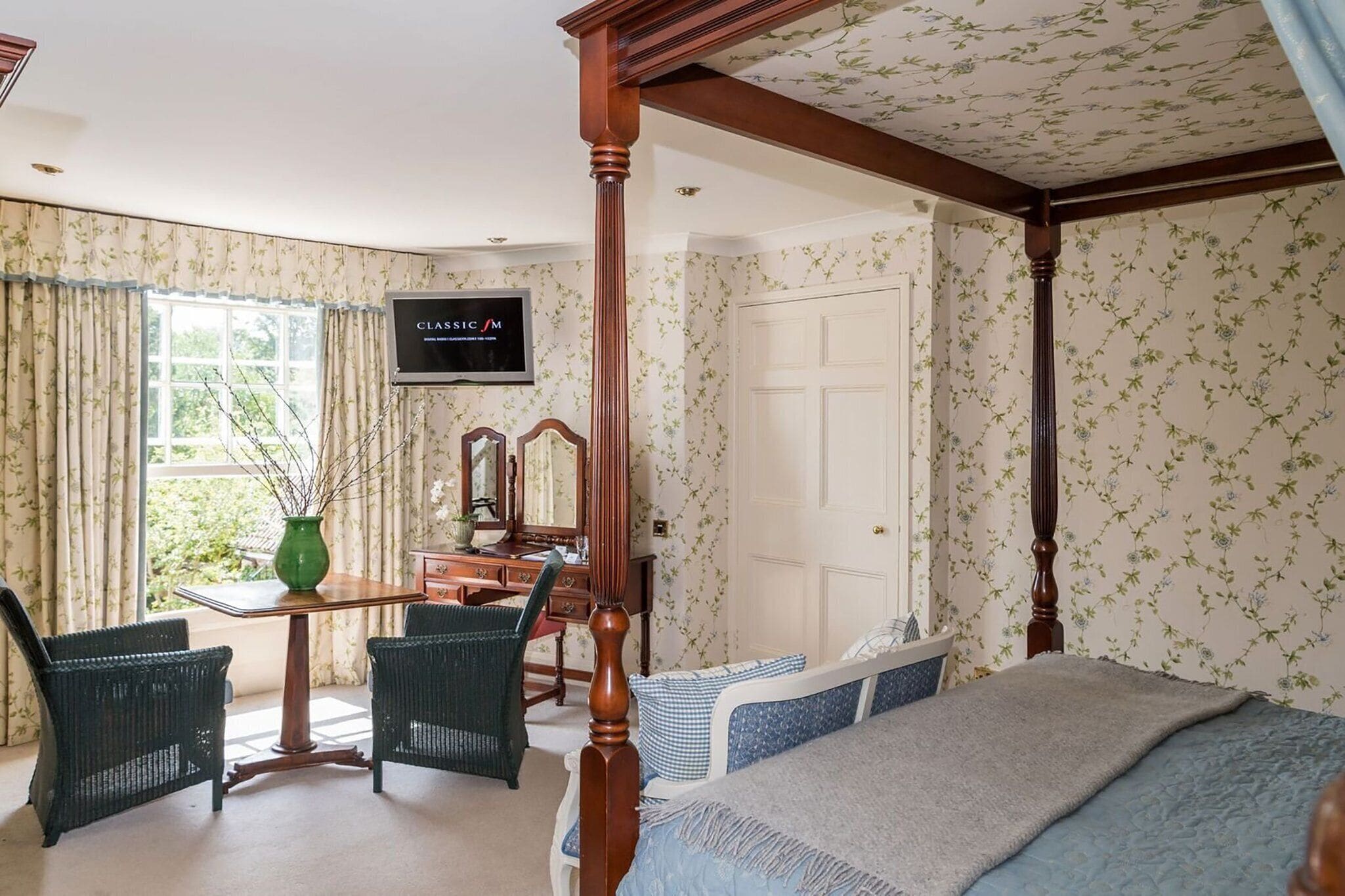 A bedroom with a four-poster bed, a desk with a mirror, and a television on the wall.