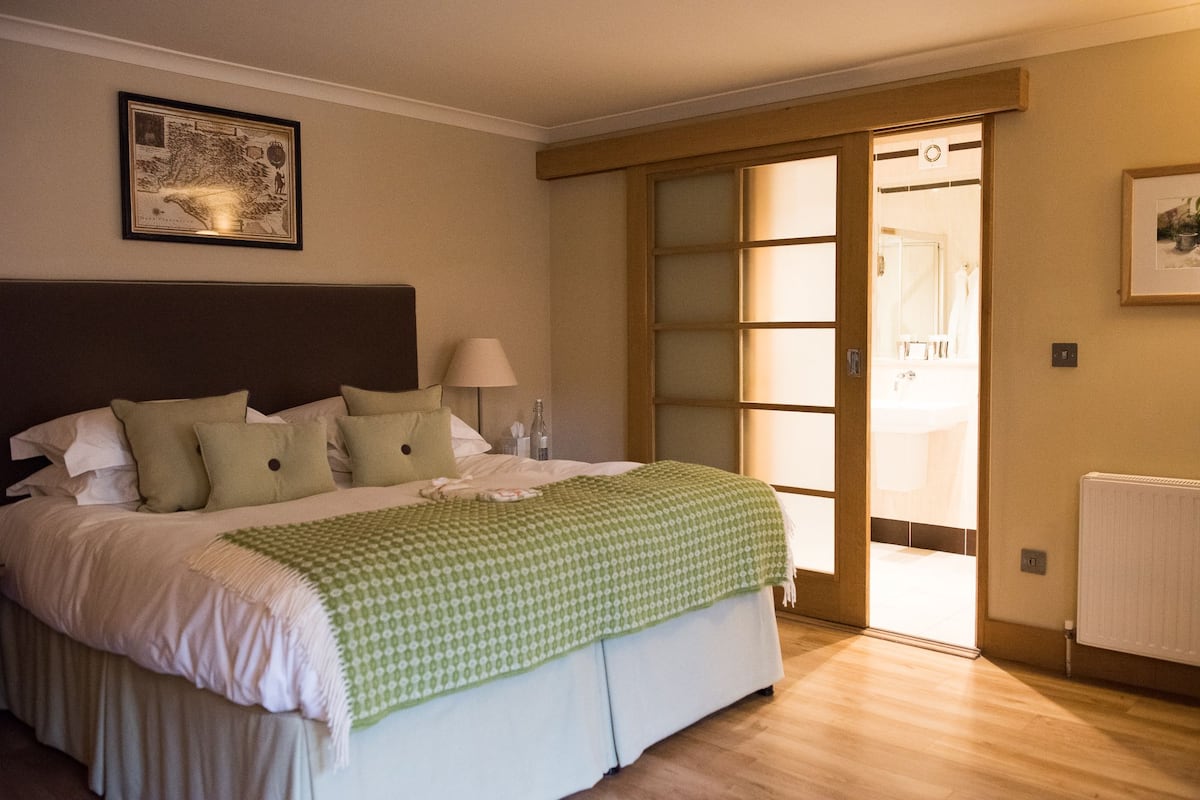A hotel room with a large bed, a nightstand, a lamp, a framed map on the wall, and a sliding glass door leading to a bathroom.