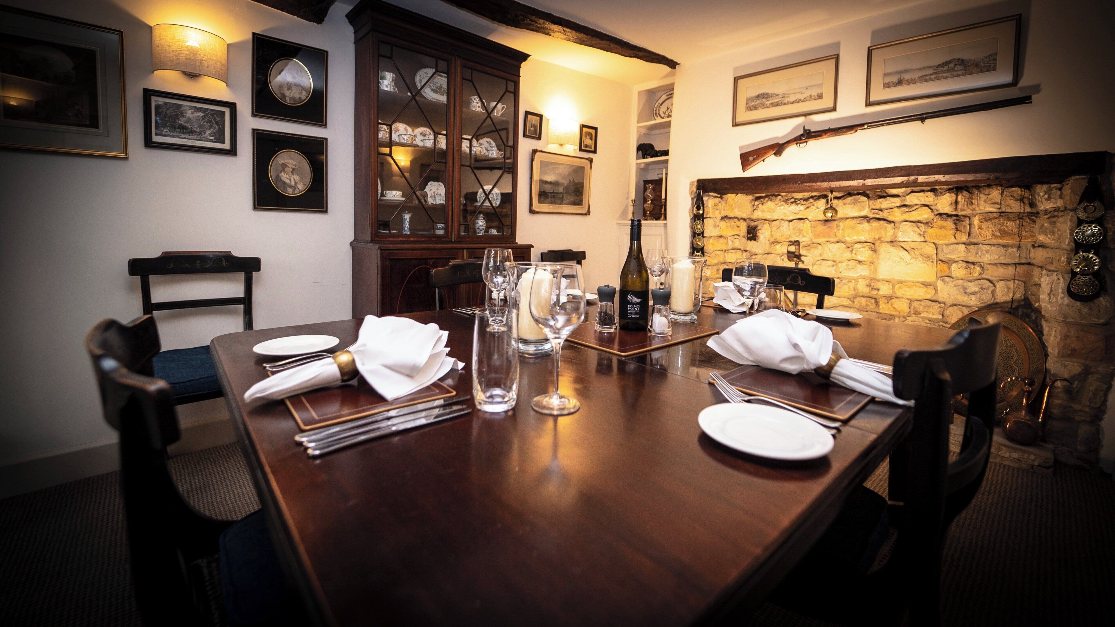 A dining room with a wooden table set for four, surrounded by chairs. There are framed pictures on the walls, a stone fireplace, and a cabinet with glass doors.