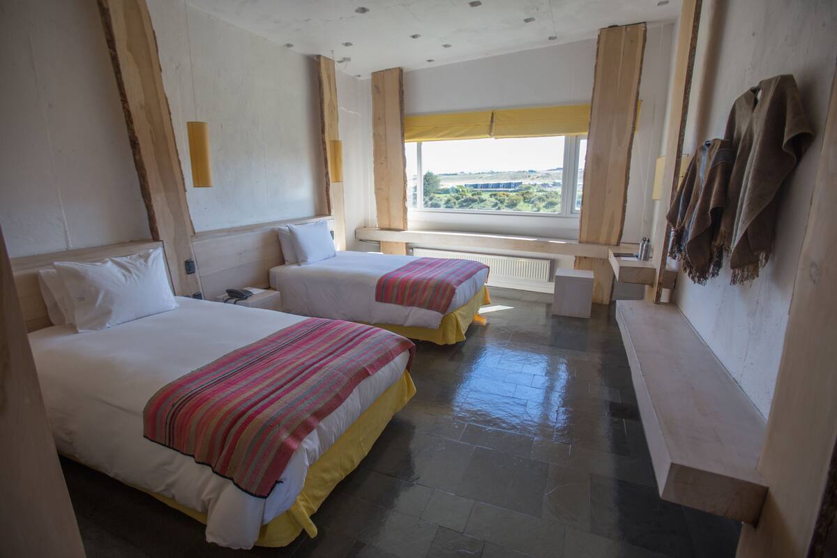 A hotel room with two beds, a window with a view, a bench, and a coat rack.