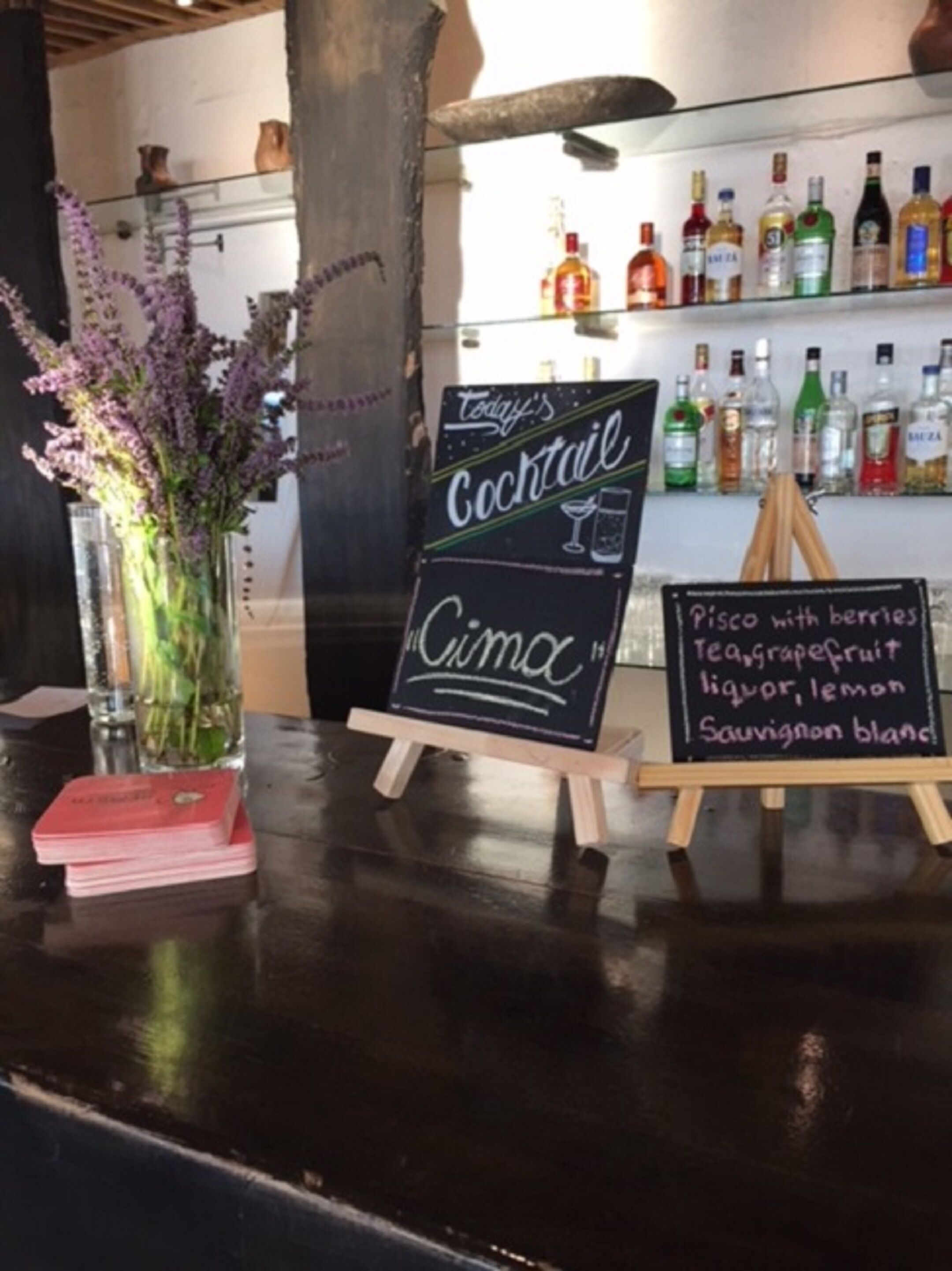 A bar area with a chalkboard menu listing a 'Cimac' cocktail and 'Pisco with berries'. There are bottles of liquor on shelves behind the bar, and a vase with purple flowers on the counter.