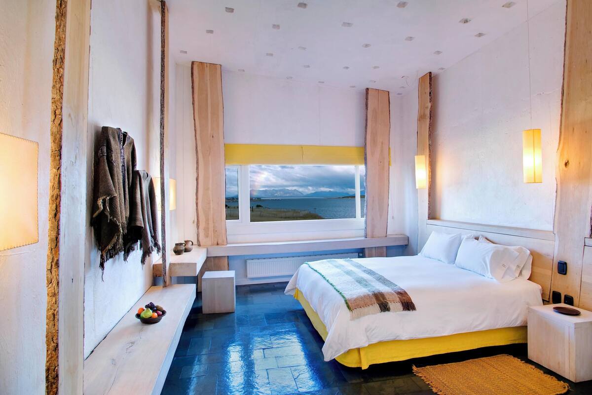 A bedroom with a bed, a window with a view, a bench, and a bowl of fruit.