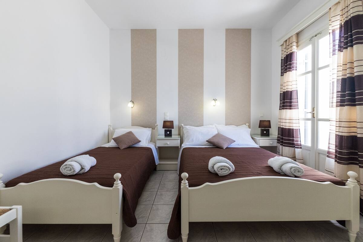 A twin bedroom with two beds, each with a brown bedspread and white headboard, a nightstand with a lamp, and a window with striped curtains.