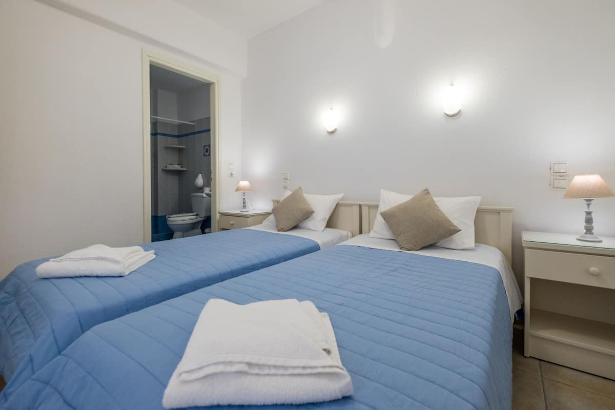 A hotel room with two beds, each with a blue quilted bedspread and white towels. There are bedside tables with lamps, a mirror, and a door leading to a bathroom.