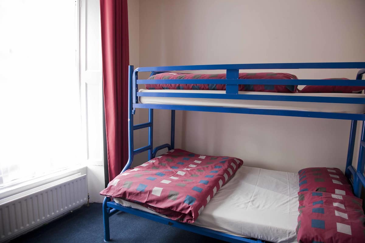 Una cama litera con una colcha roja y azul estampada, un colchón blanco y una ventana con cortina roja.