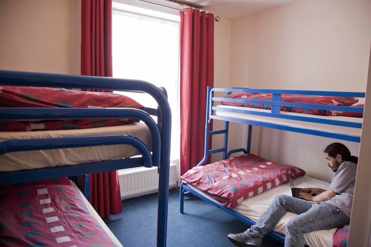 Un dormitorio con literas, una ventana con cortinas rojas y una persona sentada en una litera inferior usando una computadora portátil.