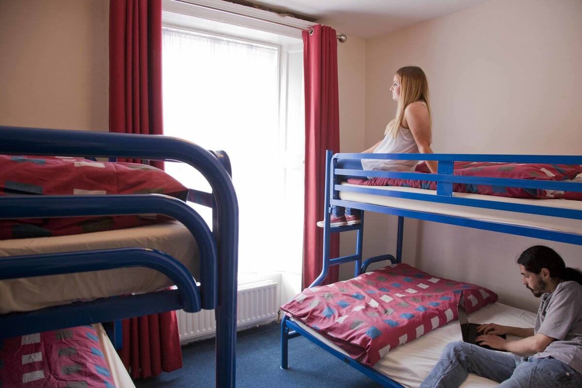 Un dormitorio con literas, una ventana con cortinas rojas y una persona usando una computadora portátil en la litera inferior.