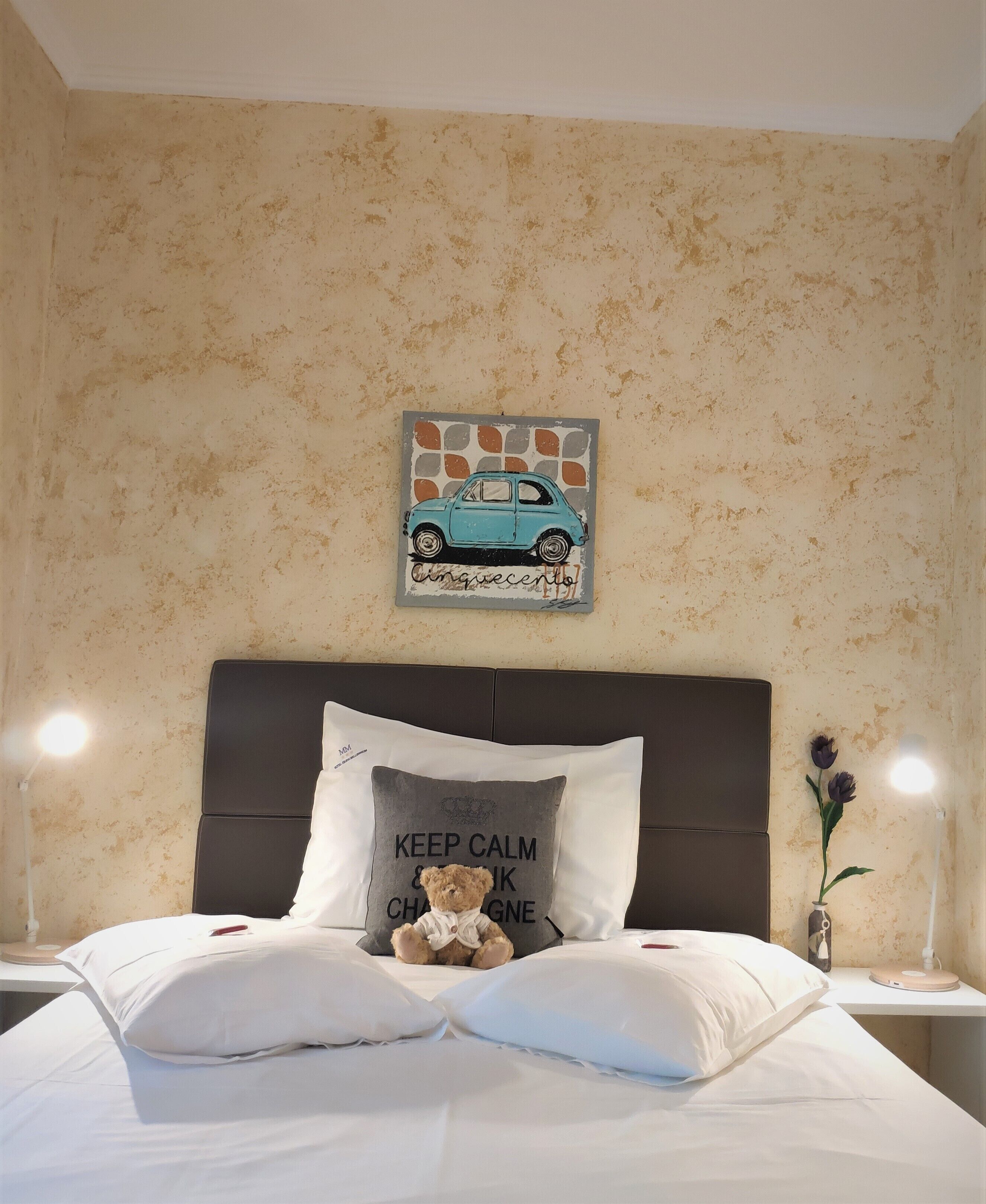 Una cama bien hecha con sábanas blancas, un almohadón con un oso y texto, una ilustración de un coche colgada en la pared y un jarrón con flores sobre una mesita de noche.