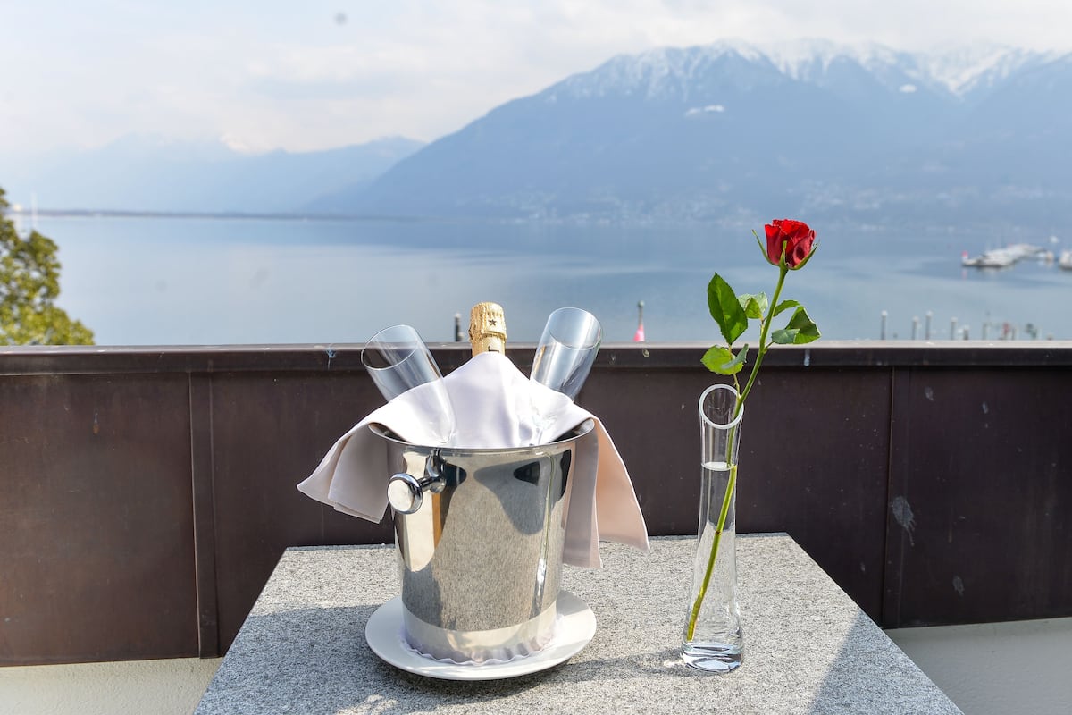 A metal bucket with a cork, a glass vase with a single rose, and a scenic view of a lake and mountains in the background.