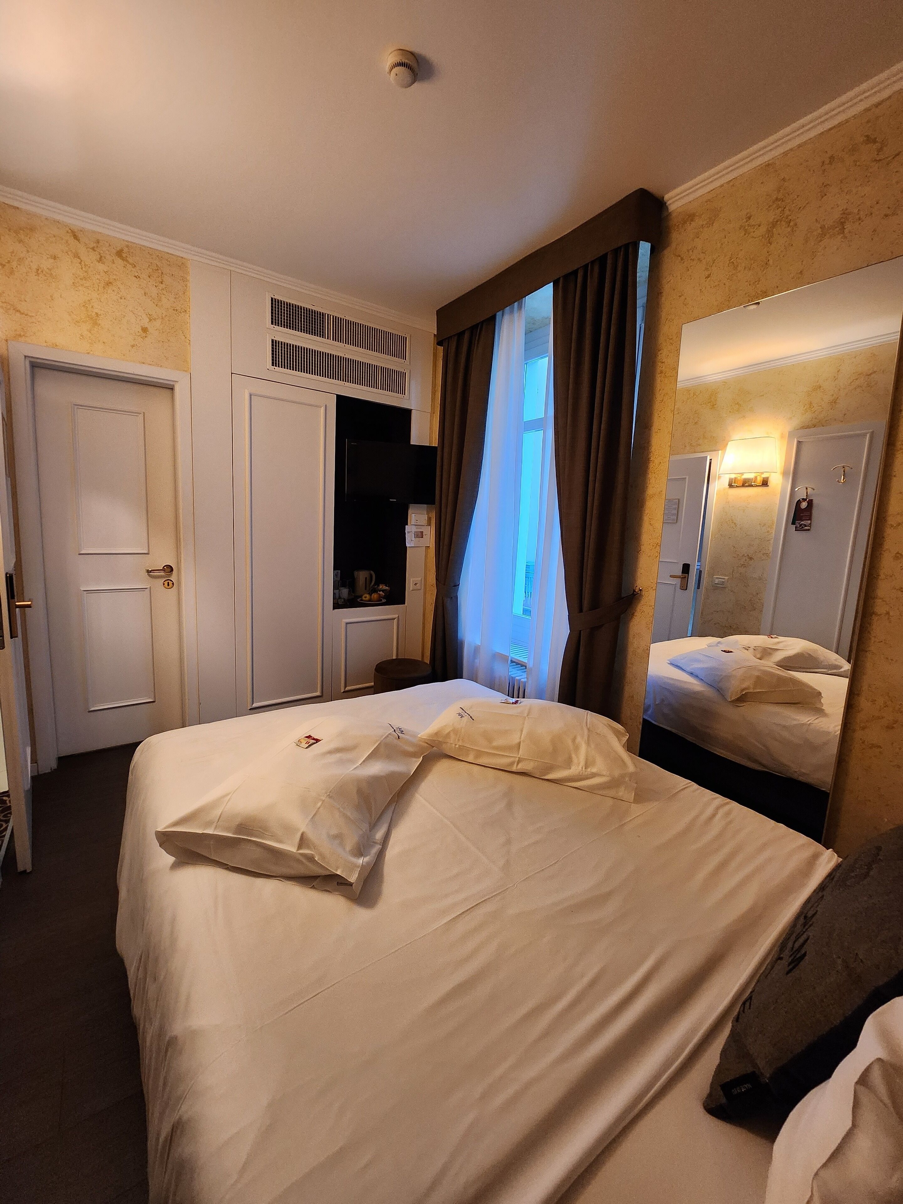 A hotel room with a large bed, a wardrobe, a mirror, and a window with curtains.