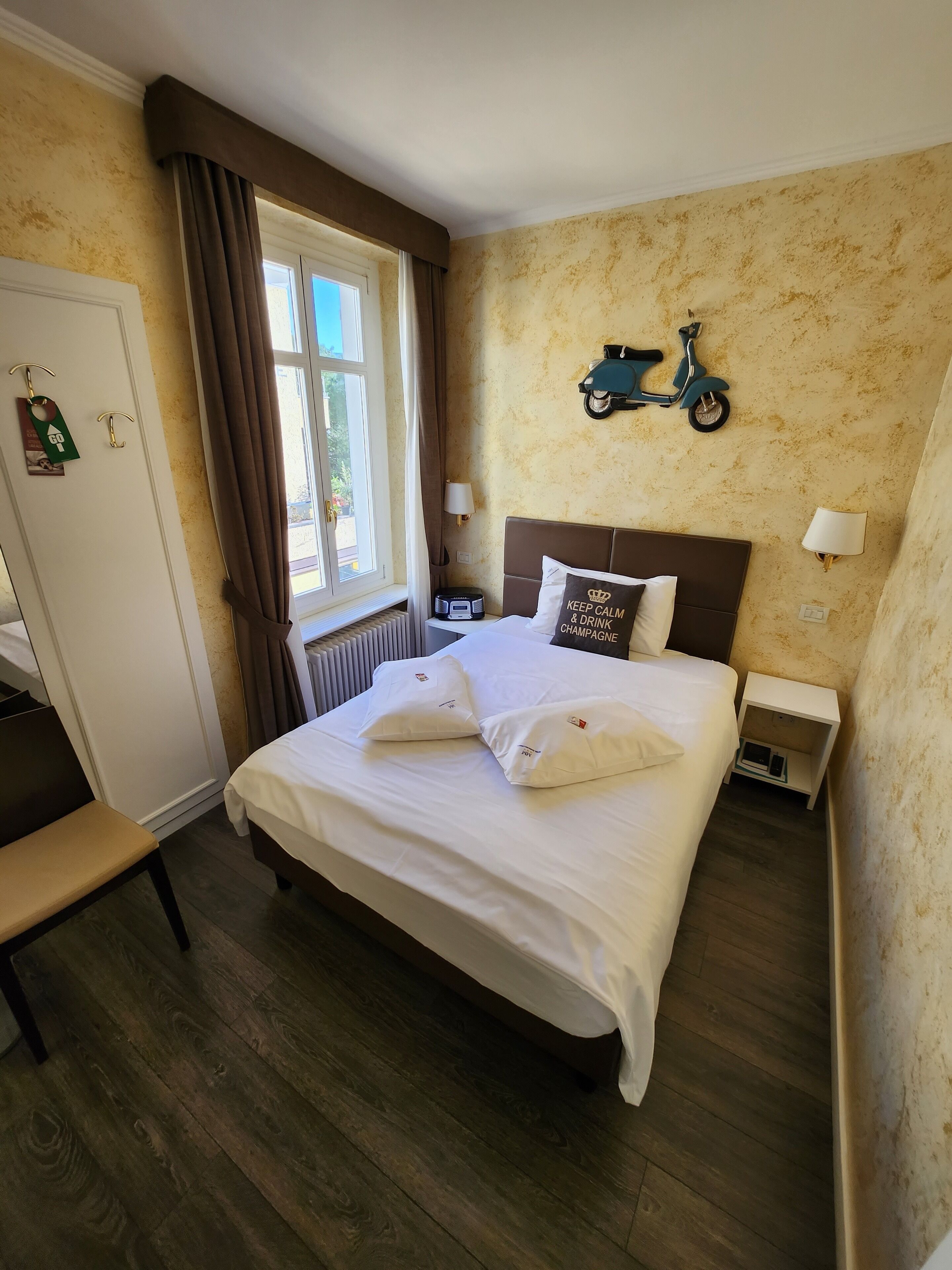 A neatly made bed with pillows, a bedside table with a lamp, a wall-mounted scooter, and a window with curtains.