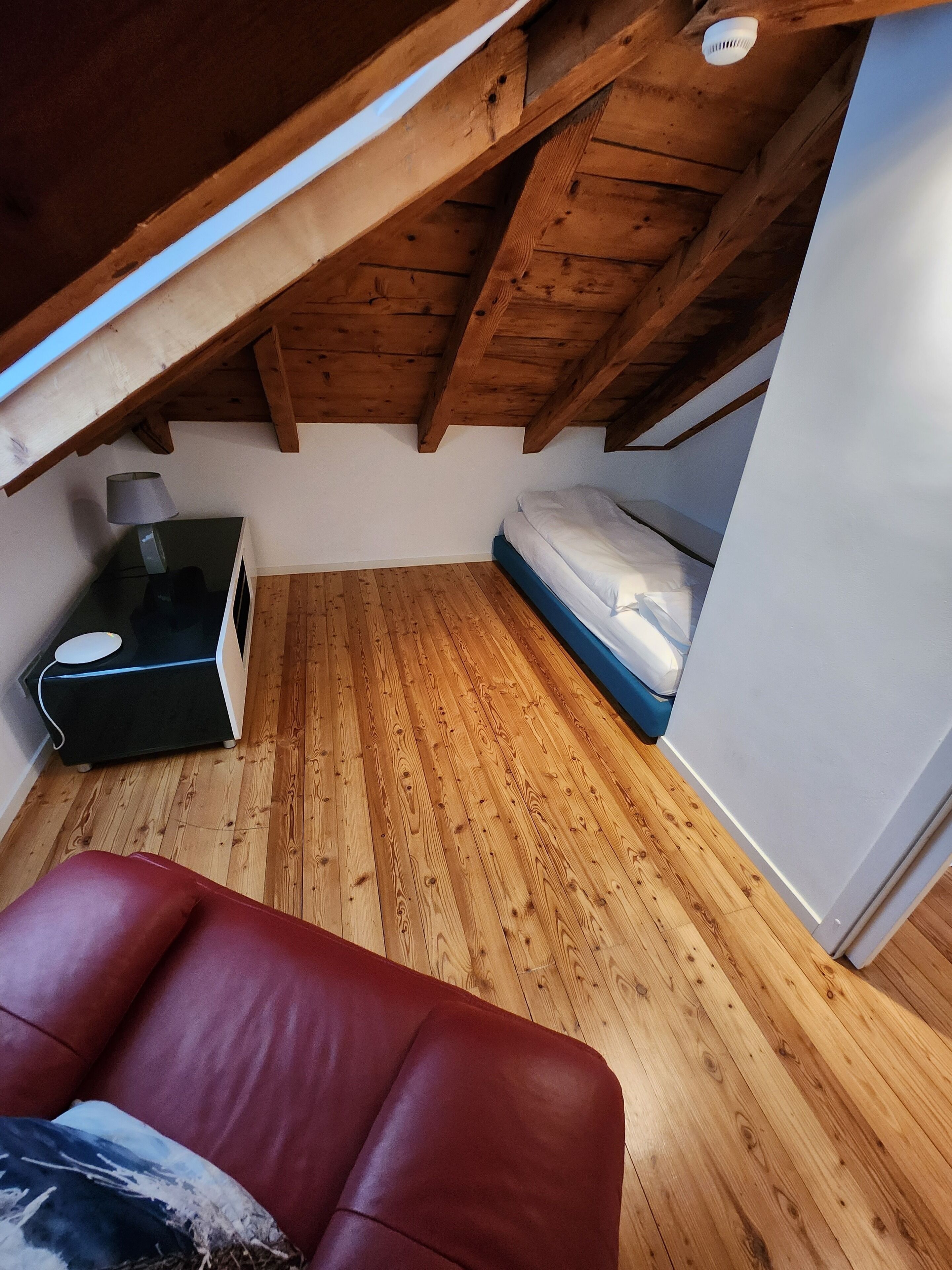 Ein kleiner, gemütlicher Dachzimmer mit Holzbalken, einem Bett, einem Schreibtisch mit Lampe und einem roten Ledersessel.