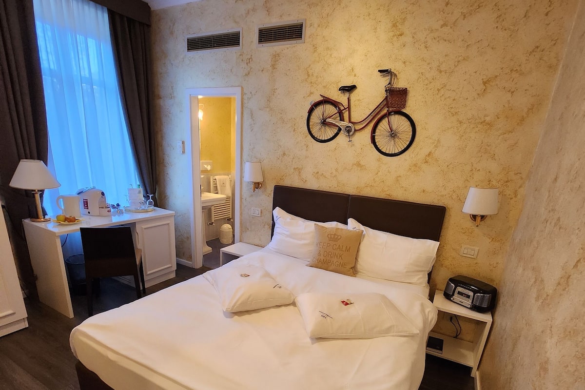 A hotel room with a large bed, a desk, a chair, a bicycle wall decoration, and a small radio on the nightstand.
