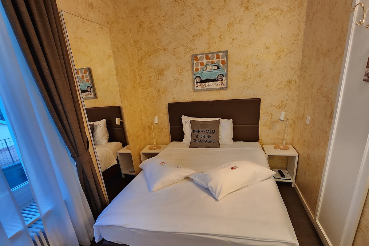 A hotel room with a large bed, two bedside tables, a nightstand, a wall clock, and a window with curtains.