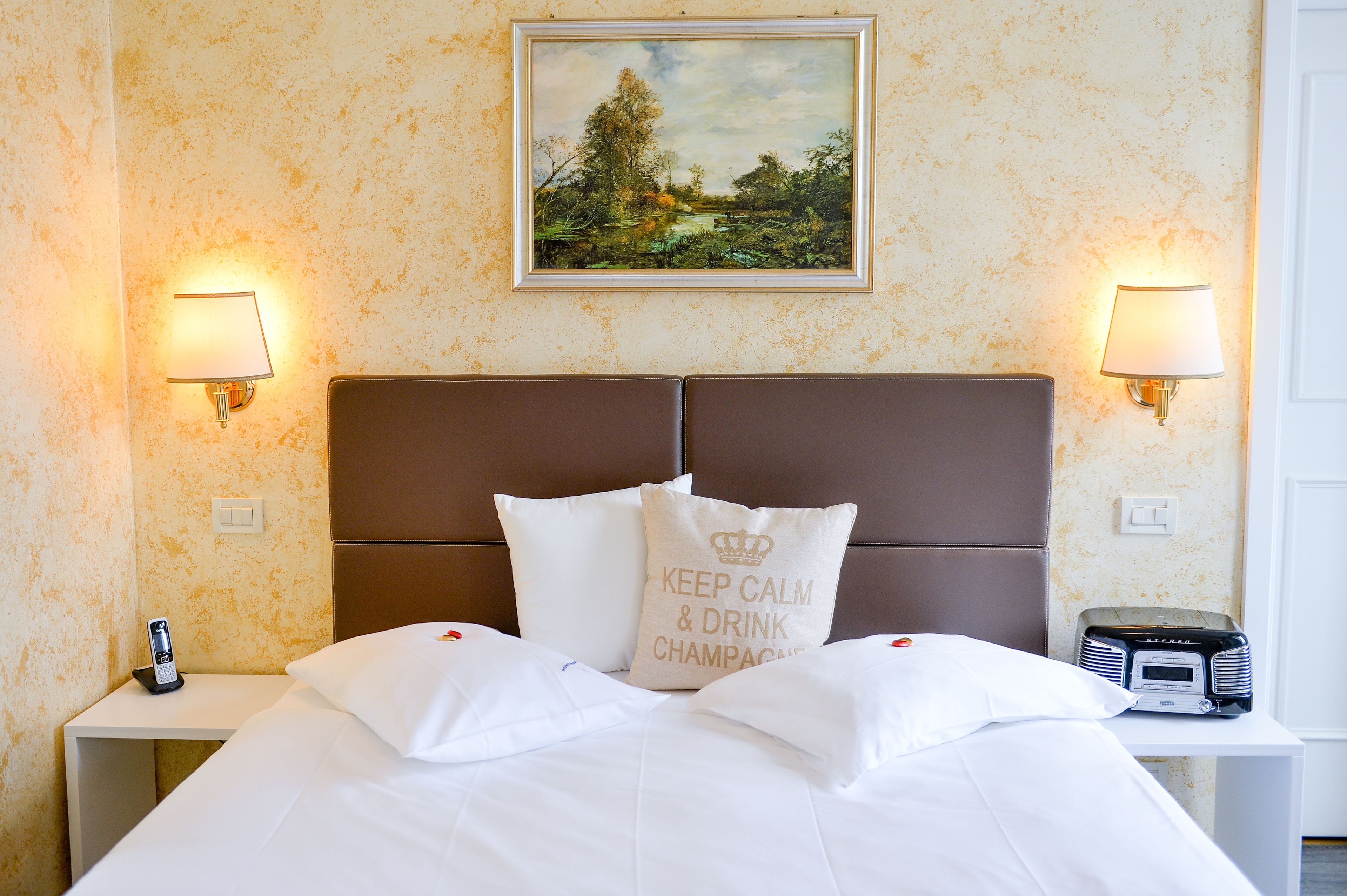 A hotel room with a bed, two bedside lamps, a painting on the wall, and a radio on the nightstand.