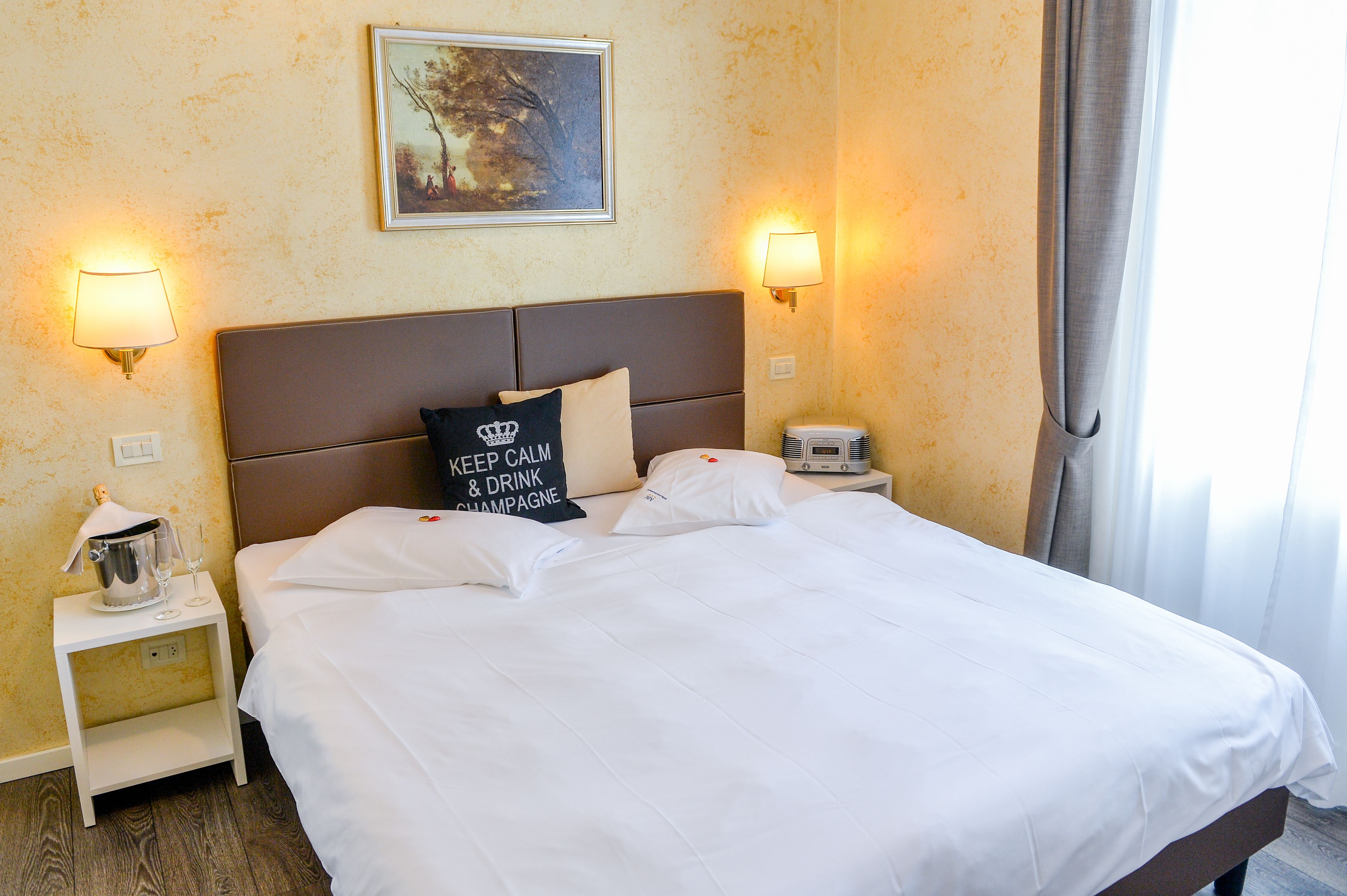 A hotel room with a large bed, two bedside lamps, a nightstand with a lamp and a radio, a framed picture on the wall, and a window with curtains.
