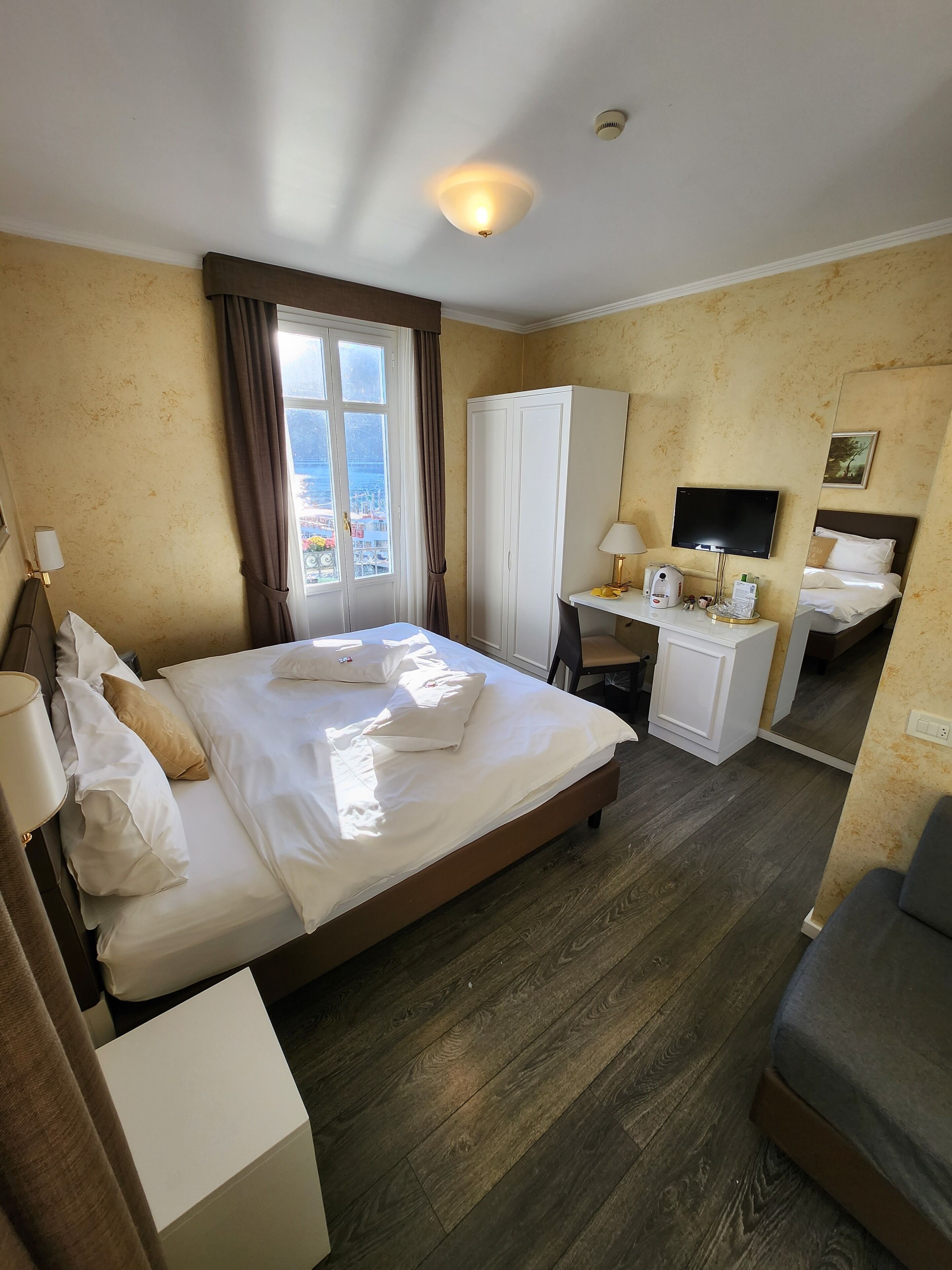 A hotel room with a bed, a desk with a chair, a wardrobe, a television, and a window with curtains.