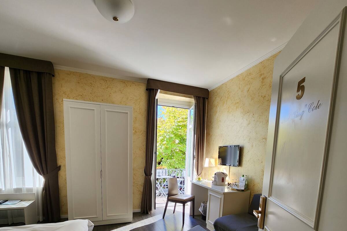 A hotel room with a bed, a desk with a chair, a small table with a lamp, a sofa, and a balcony with a chair.