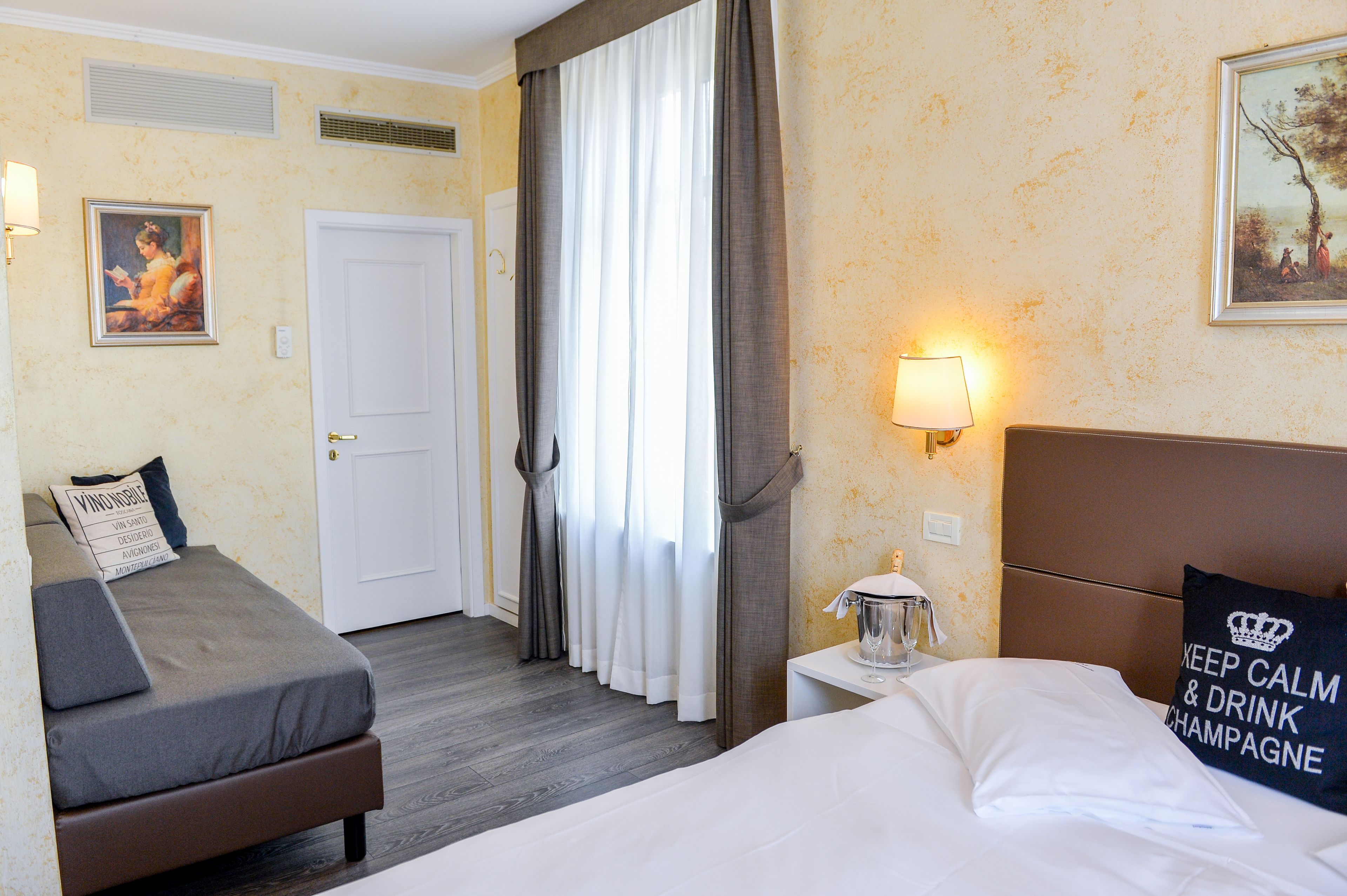 A hotel room with a bed, a sofa bed, a bedside table with a lamp, a painting on the wall, and a door.