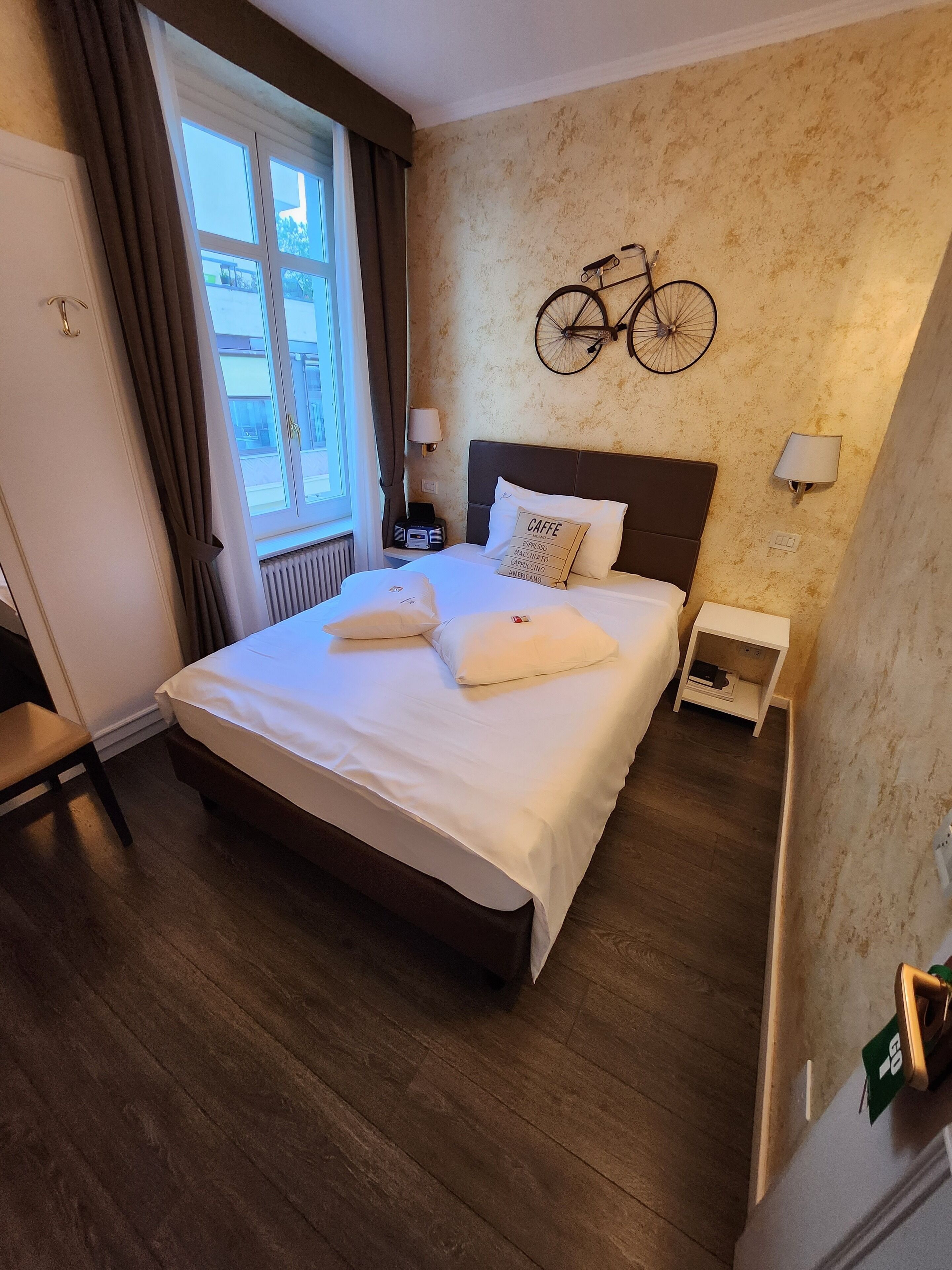 Une chambre d’hôtel avec un grand lit, une décoration murale représentant un vélo, une fenêtre avec des rideaux et une table de chevet avec une lampe.