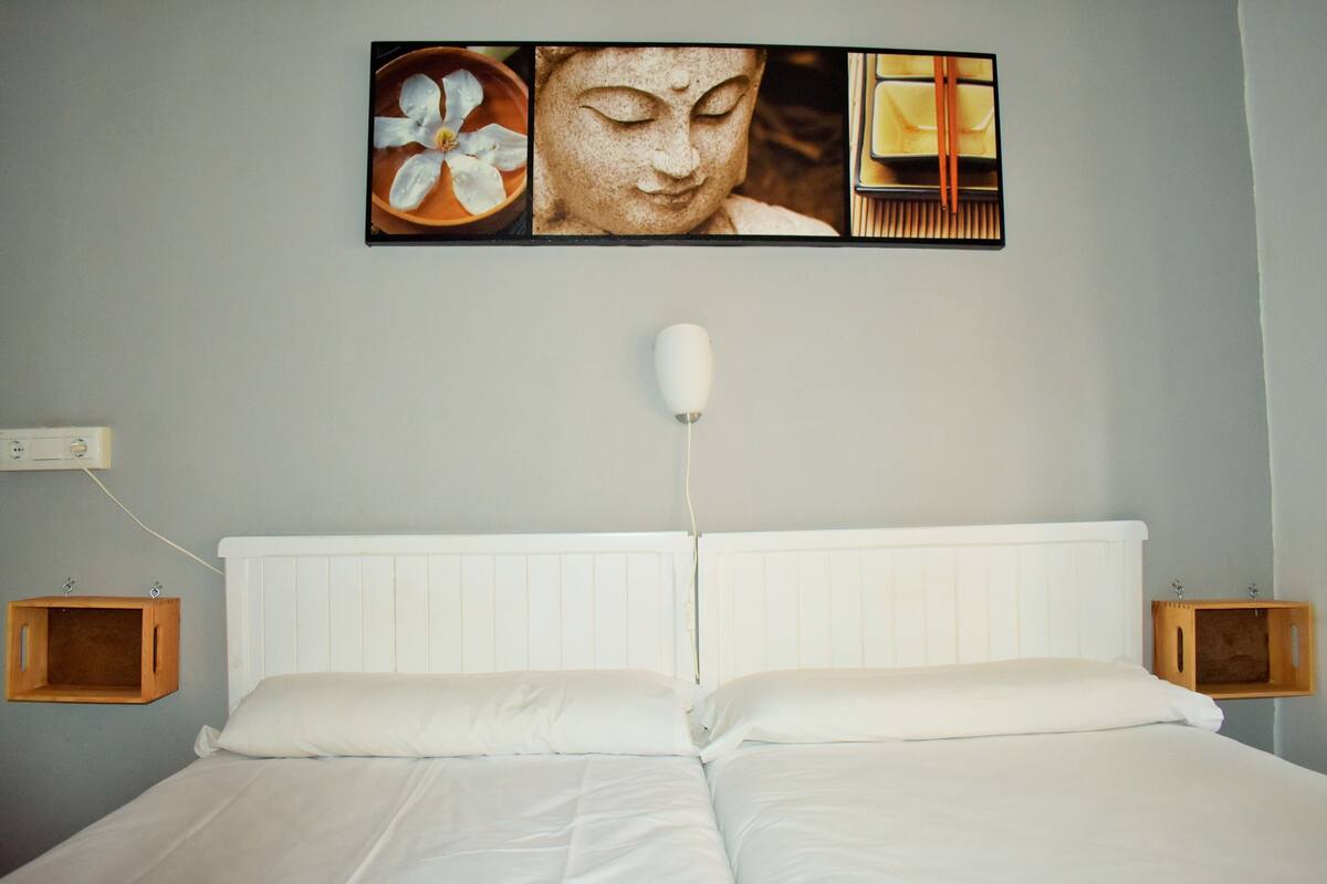 A neatly made double bed with white linens, a white headboard, and a wall-mounted light fixture. There are two wooden nightstands with a small wooden box on each.