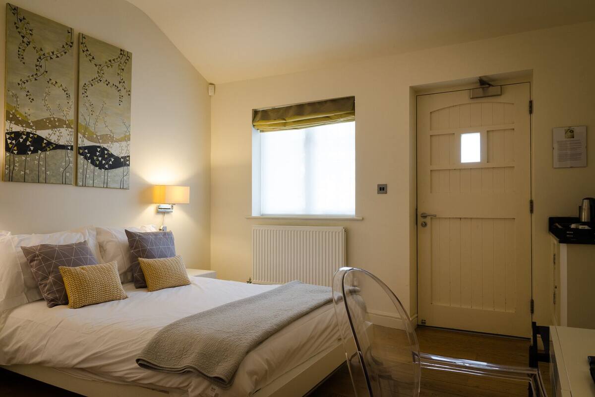 A bedroom with a bed, pillows, a bedside lamp, a window, a radiator, a door, and a chair.