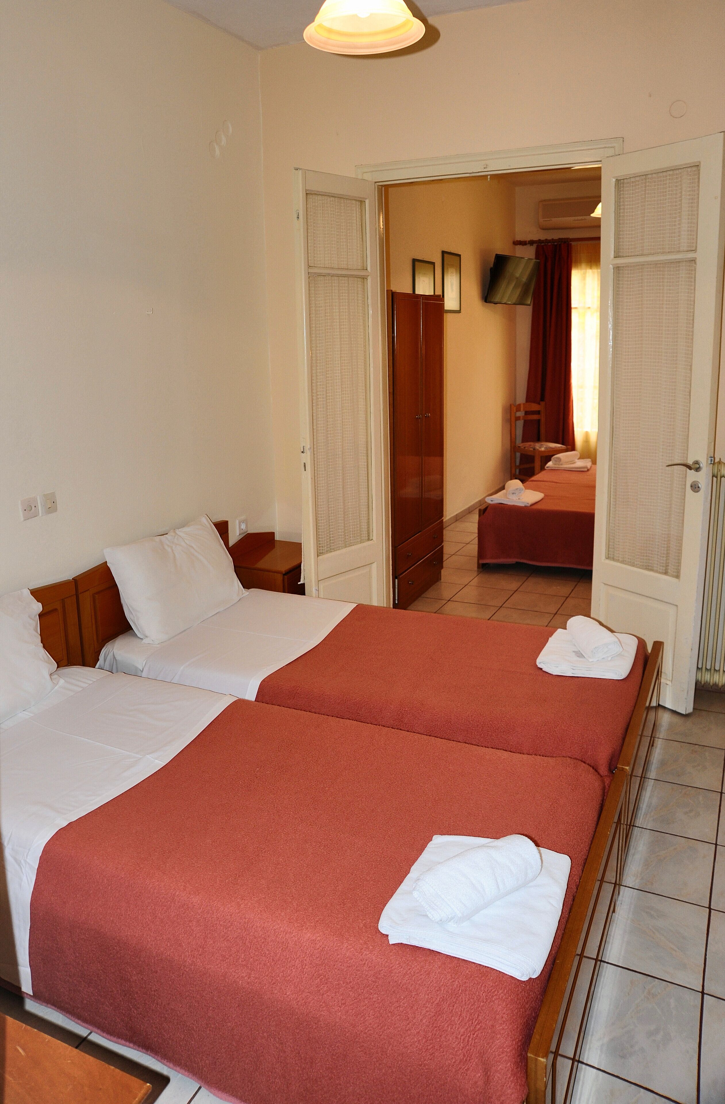 A hotel room with two beds, each with a red bedspread and white pillows. There are two nightstands, a wooden wardrobe, and a television on the wall.