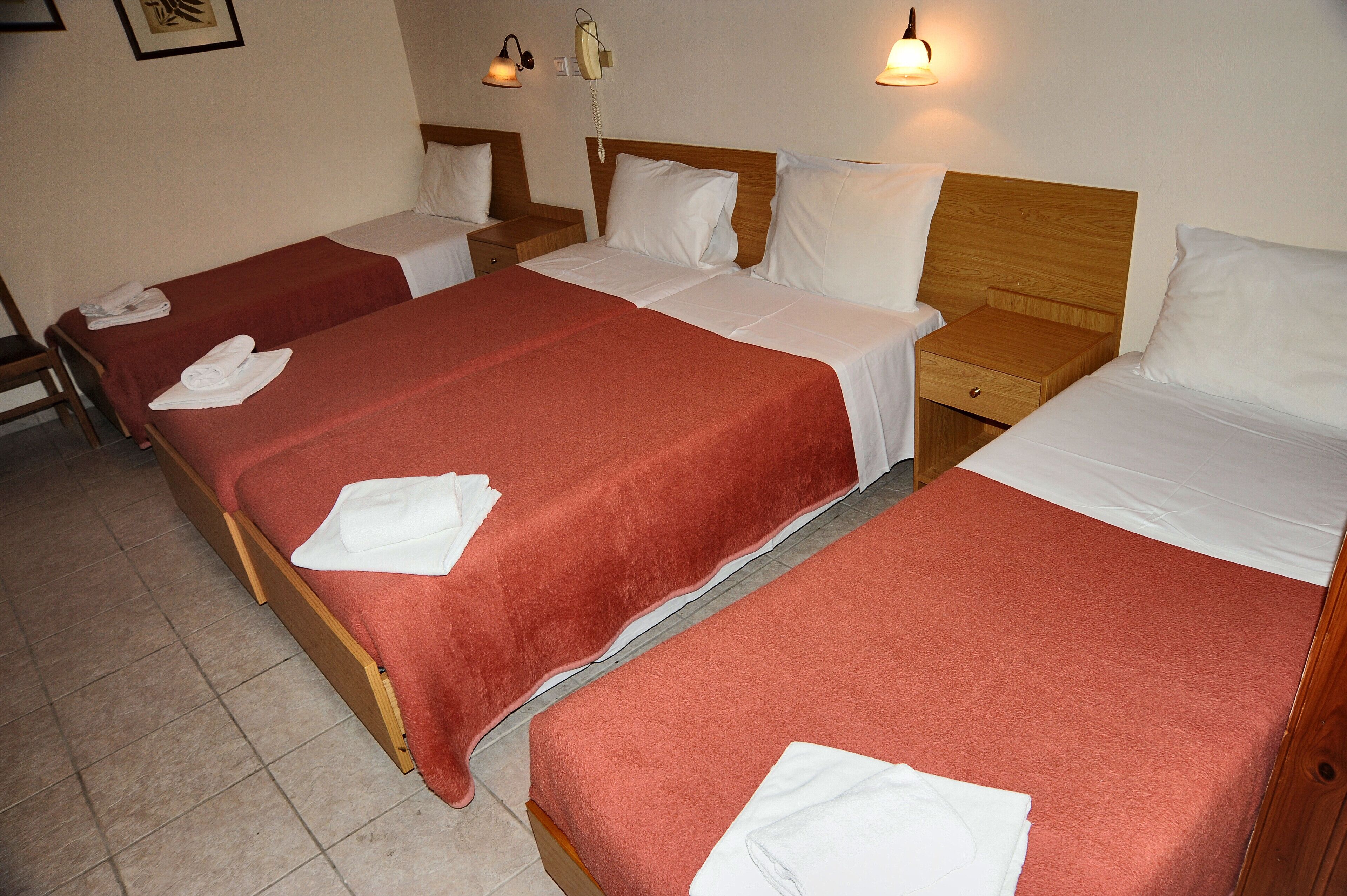 Habitació d'hotel amb tres llits individuals, cada un amb un llençol vermell i almoades blanques, i una tauleta de nit de fusta amb una làmpada.