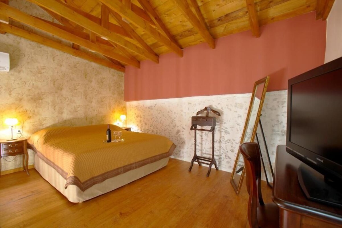A bedroom with a wooden ceiling, a bed with a brown blanket, a bedside table with a lamp, a small wooden cabinet, and a television on a stand.