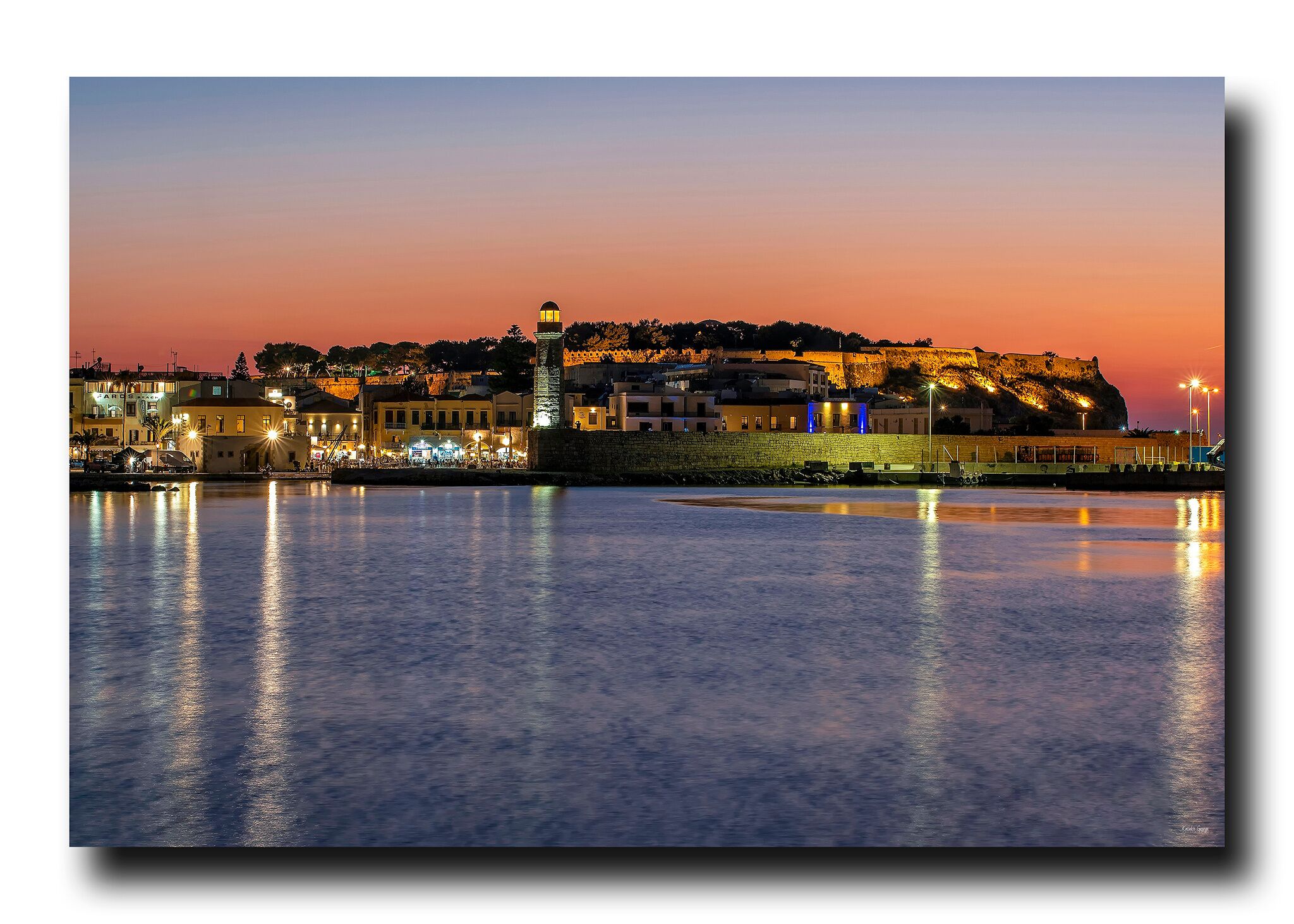 A waterfront cityscape at dusk with illuminated buildings and a prominent tower.