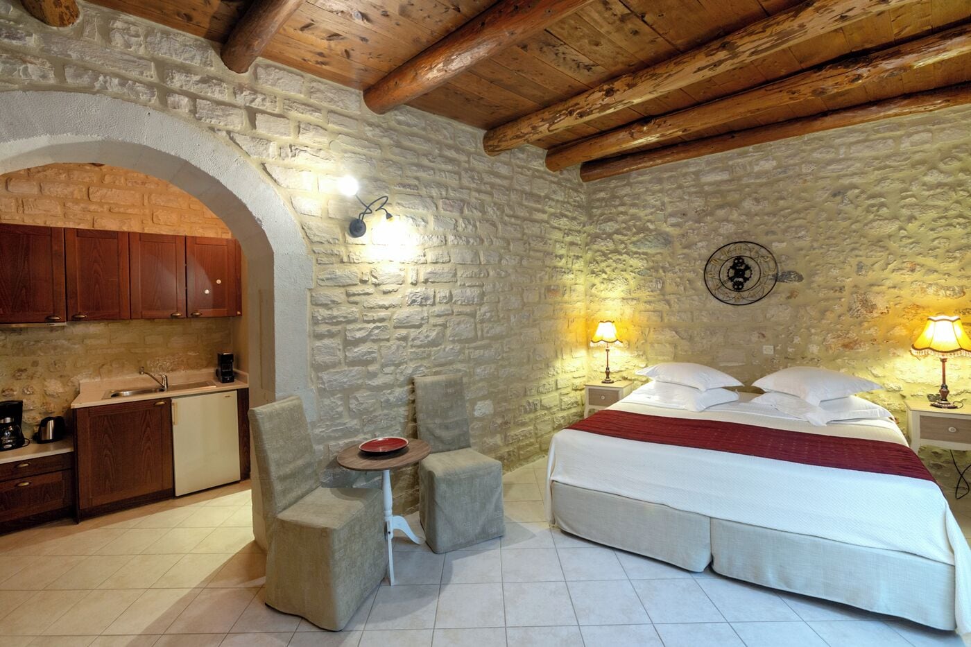 A cozy room with a stone wall, wooden ceiling beams, a bed with white linens, a small round table, and a kitchenette with wooden cabinets.