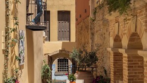 A courtyard with a table, potted plants, and a person on a balcony. - Hotel Casa Moazzo (Rethymno)