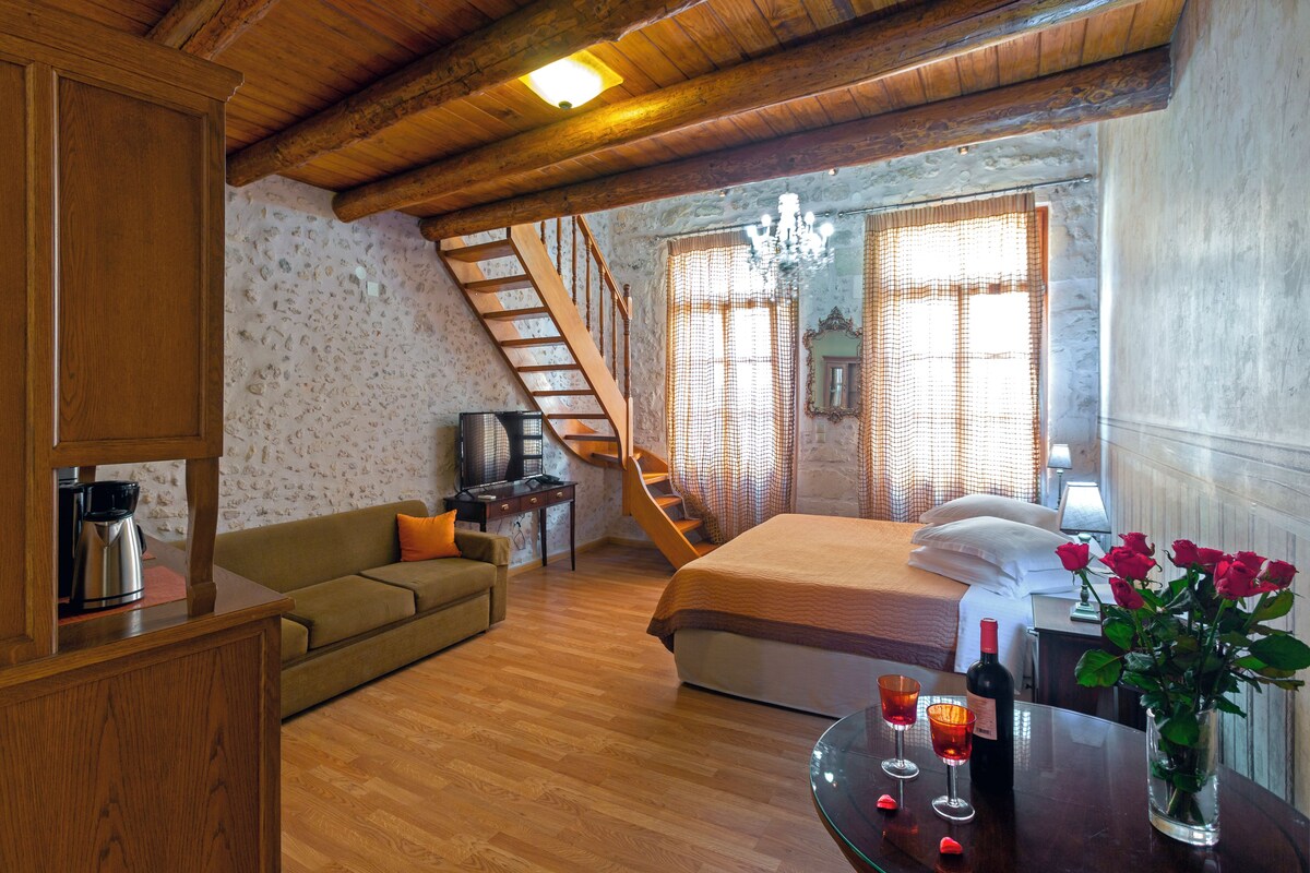 A hotel room with a wooden staircase, a bed with white linens, a wooden coffee table with glasses and a bottle, a sofa, and a wall-mounted TV.