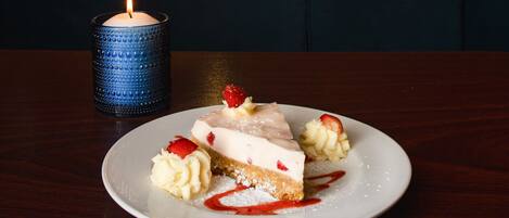 Un plato de postre con una rebanada de pastel de queso, fresas y crema batida sobre una mesa de madera.
