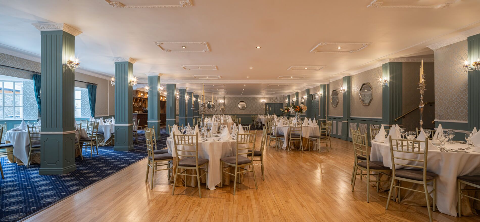 A spacious dining hall with multiple tables and chairs, elegant chandeliers, and a patterned carpet.