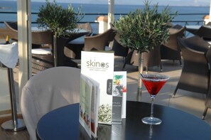 A menu and a red cocktail on a table at a seaside restaurant with wicker chairs and potted plants.