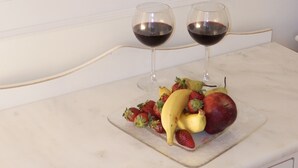 Habitación de hotel con cama, dos mesitas de noche, un tocador, un espejo y una encimera de mármol con frutas y copas de vino.