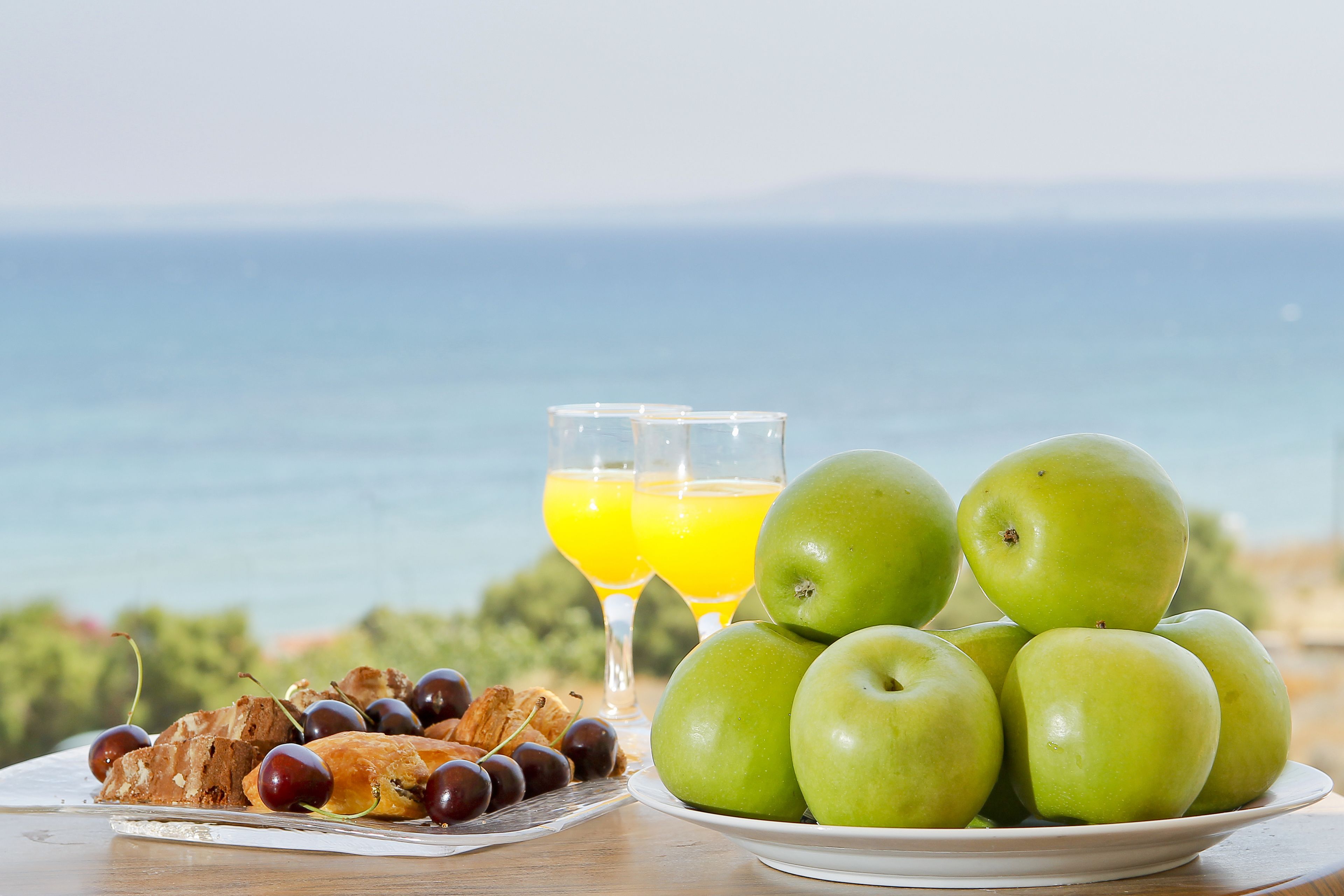 A table with a plate of green apples, a glass of orange juice, and a plate of pastries and cherries.