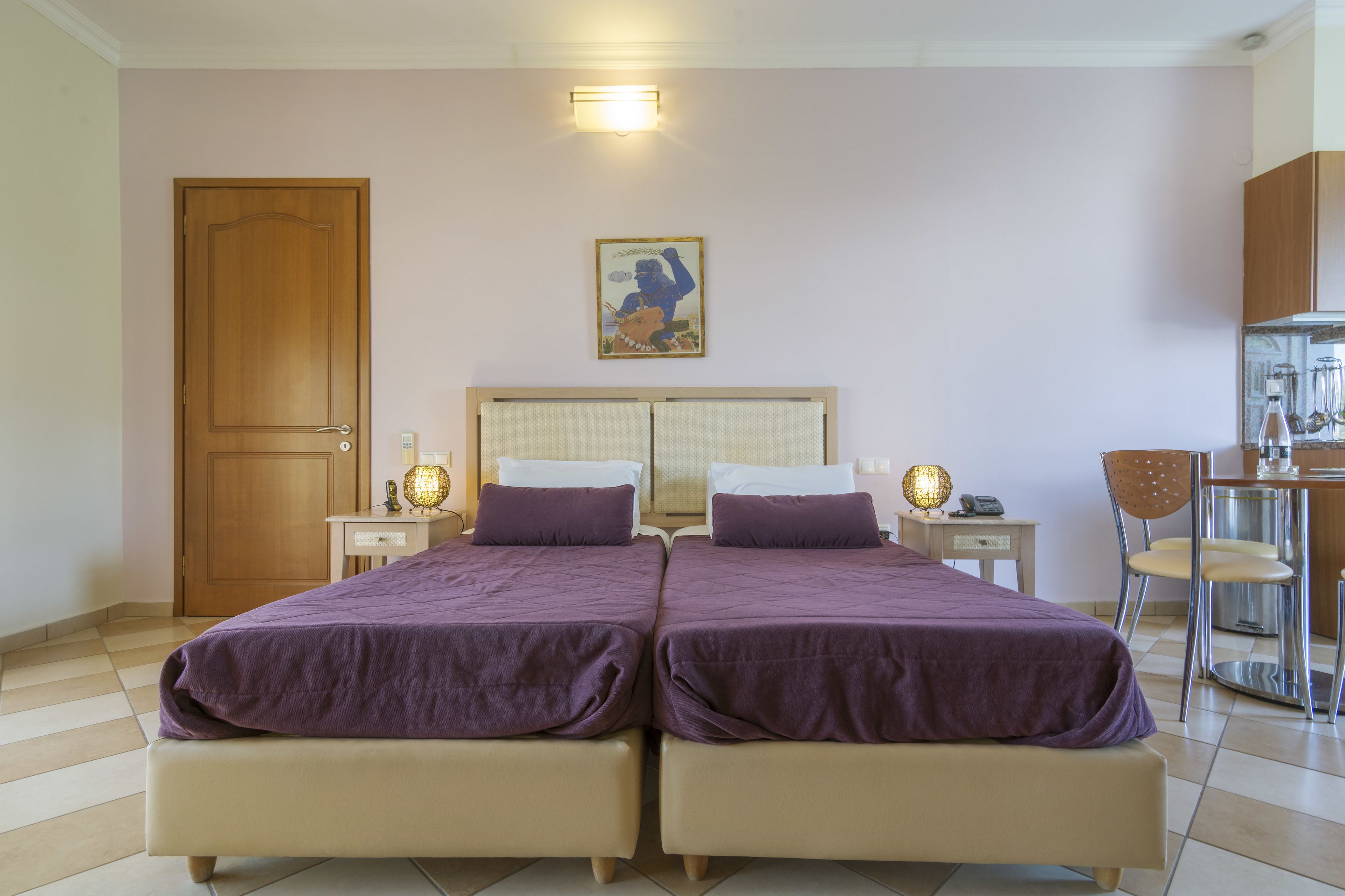 Una cama doble con cabecera, dos mesitas de noche con lámparas, una puerta de madera, un cuadro enmarcado en la pared y una zona de kitchenette con una silla.