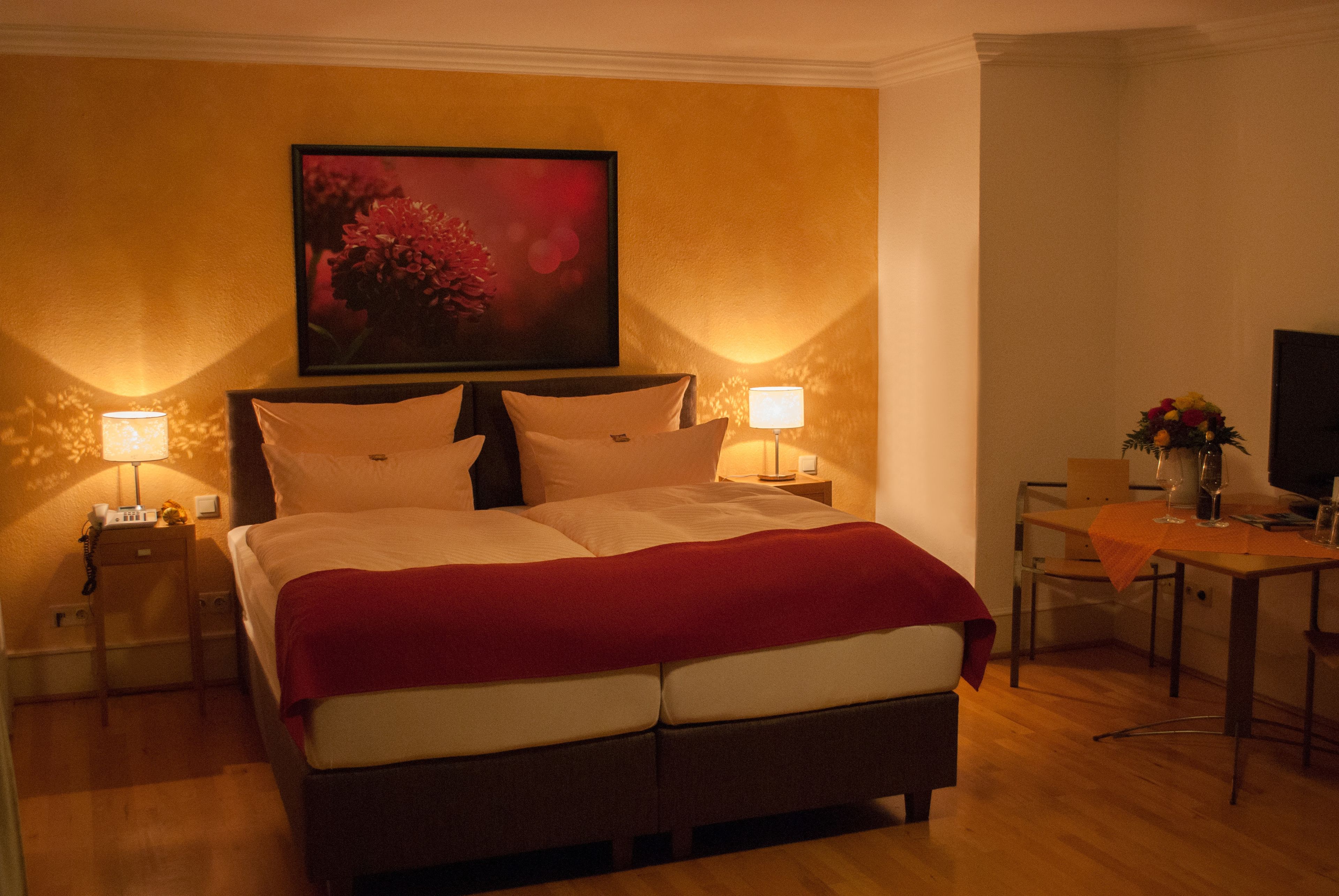 Habitación de hotel con una cama grande, dos mesitas de noche con lámparas, una mesita pequeña con un florero y un cuadro en la pared.
