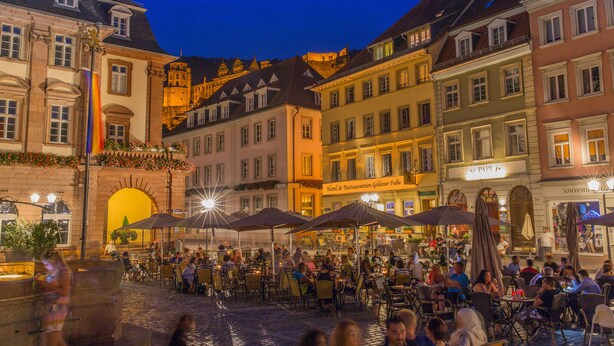 Un animado café al aire libre con mesas y sombrillas, rodeado de edificios históricos con diversos estilos arquitectónicos.