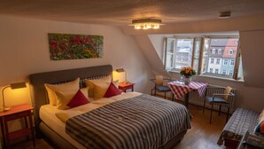 Un dormitorio con una cama grande, mesitas de noche con lámparas, una pequeña mesa con un florero y vistas a edificios a través de una ventana.