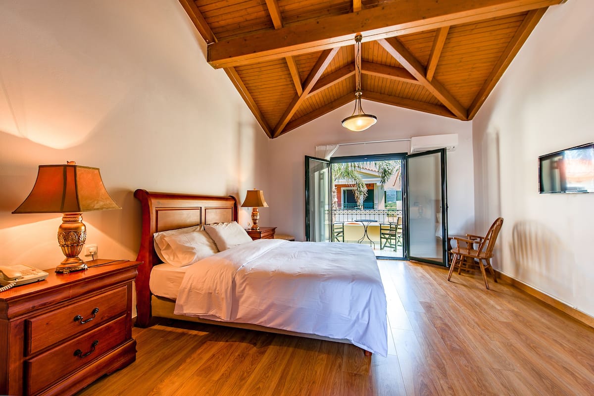Ein Schlafzimmer mit Holzbalkendecke, einem Bett mit weißen Leinen, einem Nachttisch mit Lampe und Blick auf einen Außenbalkon.