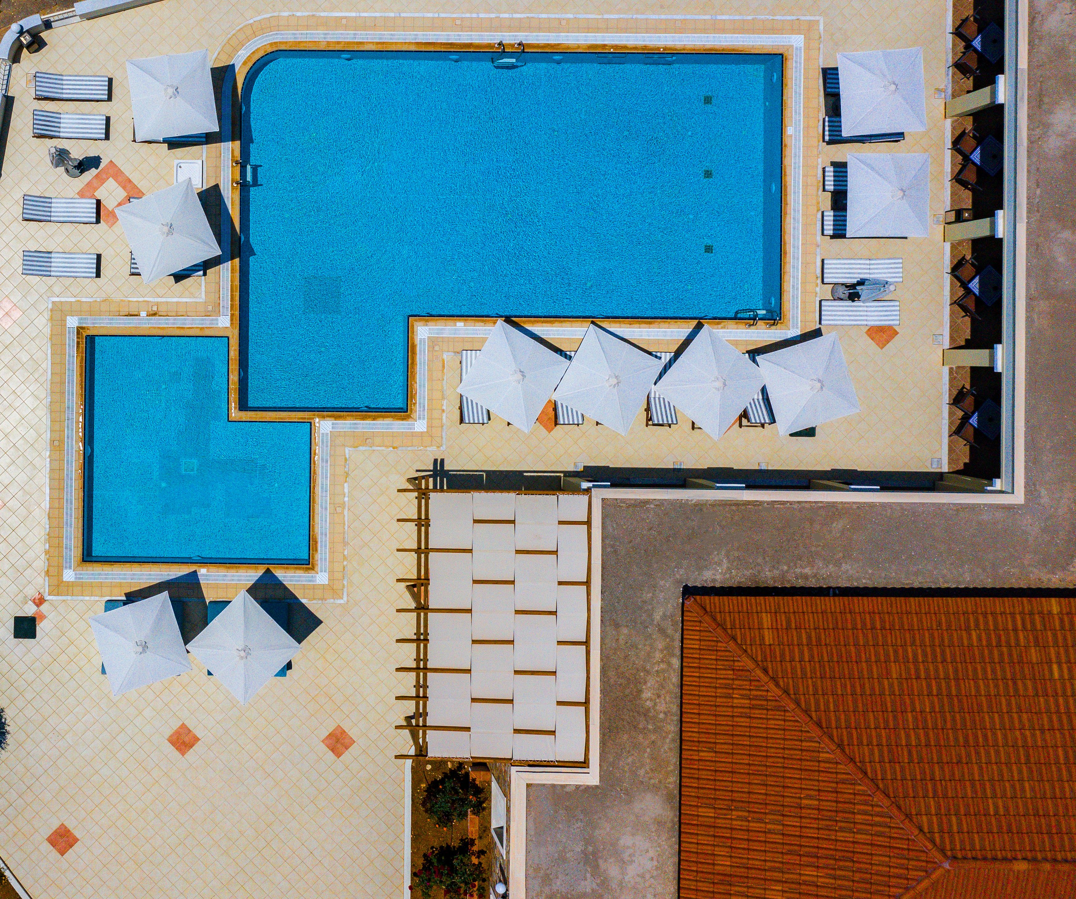 An aerial view of a hotel with two swimming pools, sun loungers, umbrellas, and a covered seating area.