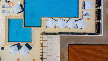 An aerial view of a hotel with two swimming pools, sun loungers, umbrellas, and a covered seating area.
